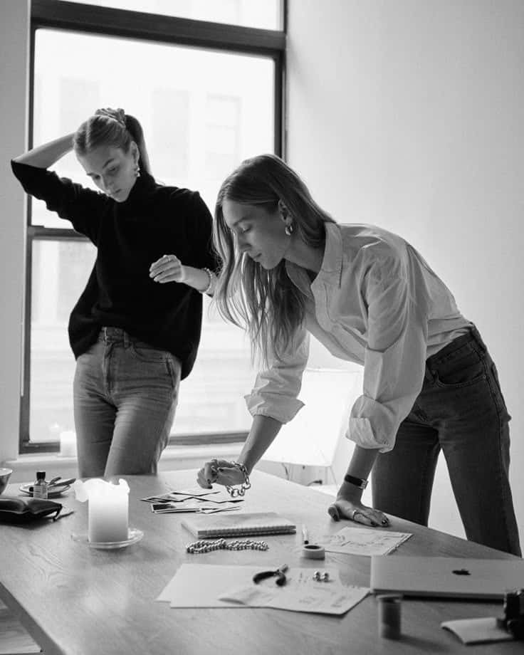 Two women collaborating on design concepts at a desk with papers, photos, and jewelry, in a sunlit office space.