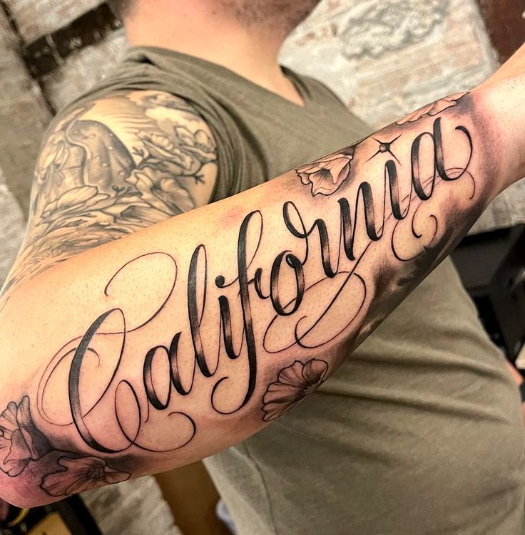 Arm tattoo with California in elegant script, surrounded by floral designs on a person wearing a grey shirt.