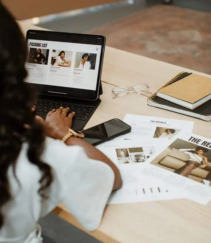 Person working on a laptop with a packing list on screen, surrounded by documents, phone, glasses, and notebooks on a wooden table.