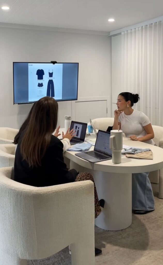 Two women in a meeting room discussing fashion design sketches displayed on a screen, laptops open on the table.