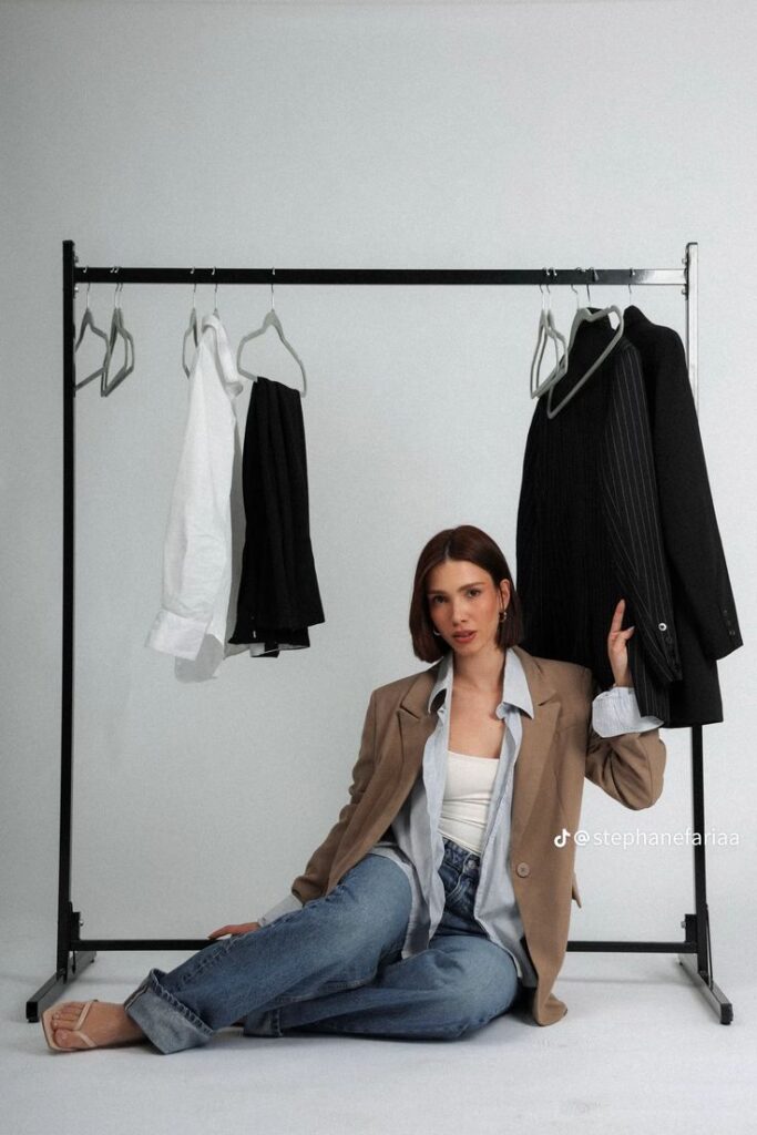 Woman sitting on floor in casual outfit with clothes rack, featuring jackets and shirts; minimalist wardrobe fashion concept.