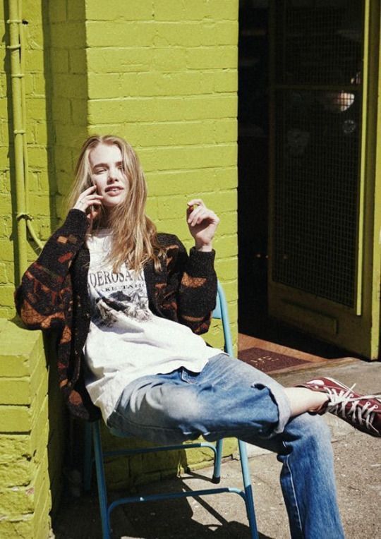 _ 19 Person in casual outfit sits on a chair against a lime green brick wall, enjoying a phone call. Sunlit, urban setting with relaxed atmosphere. | Sky Rye Design Person in casual outfit sits on a chair against a lime green brick wall, enjoying a phone call. Sunlit, urban setting with relaxed atmosphere.