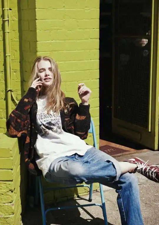Person in casual outfit sits on a chair against a lime green brick wall, enjoying a phone call. Sunlit, urban setting with relaxed atmosphere.
