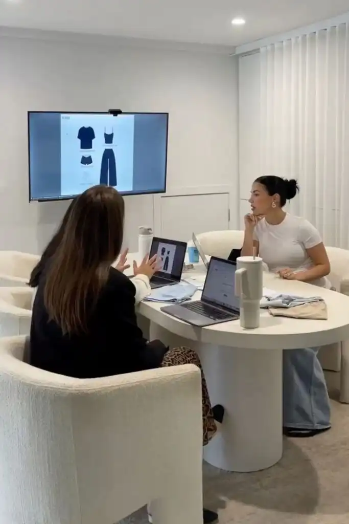 Women collaborate in a modern office setting, discussing fashion designs displayed on a screen, with laptops and fabric swatches on the table.