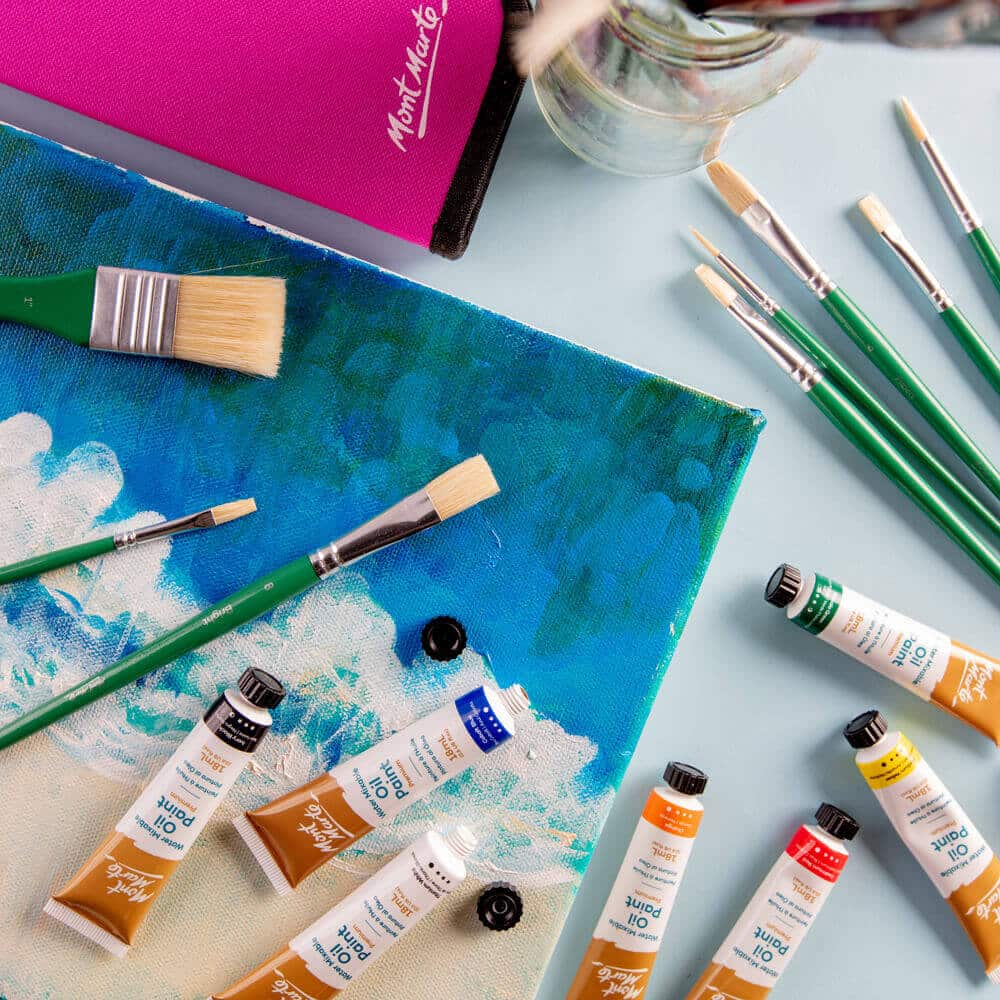 Assortment of oil paints and brushes on a painted canvas, featuring vibrant colors and art supplies from Mont Marte on a light background.