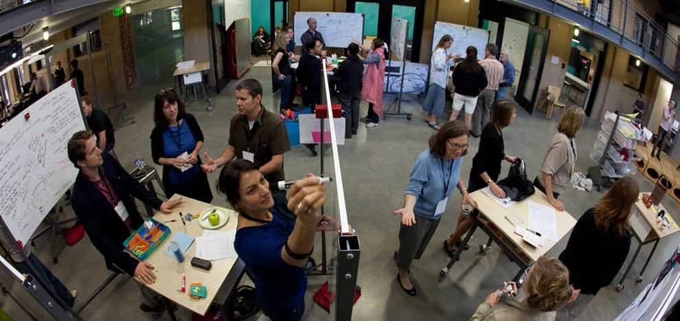 1_afAfYgGvyTMdvGe7pKqpIQ People collaborating in a creative workshop, surrounded by whiteboards and tables, inside a modern workspace. | Sky Rye Design People collaborating in a creative workshop, surrounded by whiteboards and tables, inside a modern workspace.