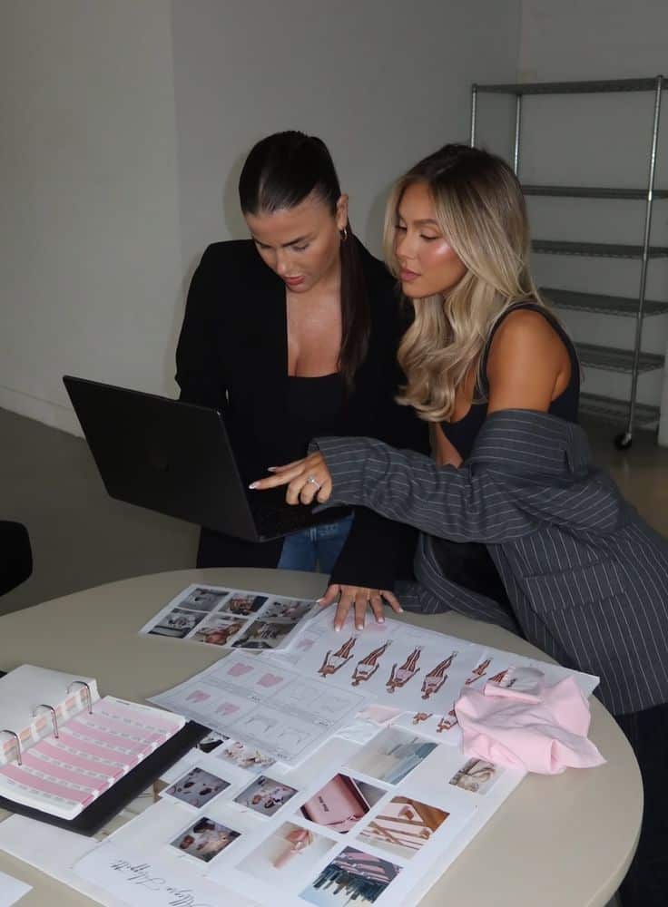 _ 2 Two women in a design meeting, reviewing a laptop and fashion sketches on a table. Office setting, collaboration in progress. | Sky Rye Design Two women in a design meeting, reviewing a laptop and fashion sketches on a table. Office setting, collaboration in progress.