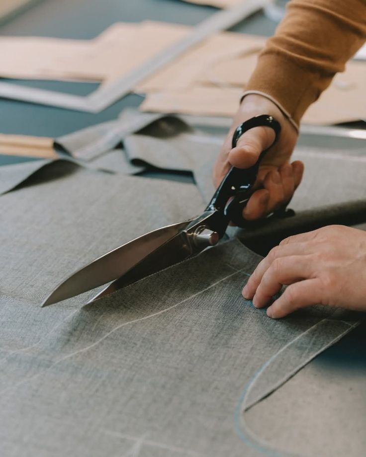 _ 2 Tailor cutting gray fabric with scissors on a pattern table, showcasing precision and craftsmanship in fashion design. | Sky Rye Design Tailor cutting gray fabric with scissors on a pattern table, showcasing precision and craftsmanship in fashion design.