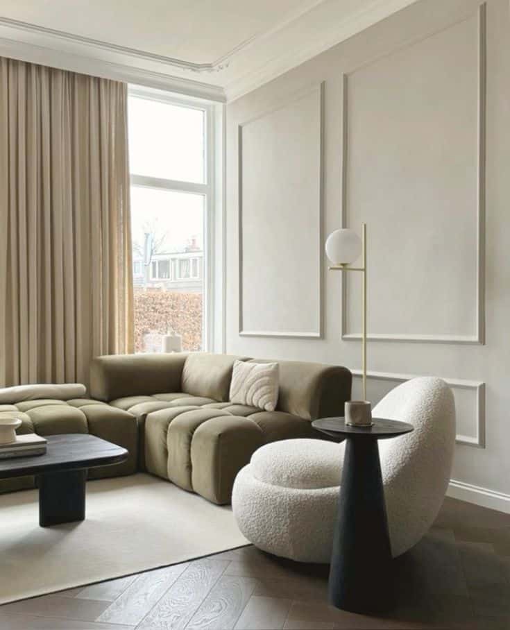Modern living room with a plush olive green sectional sofa, beige curtains, round white chair, and sleek black table. Bright window light.