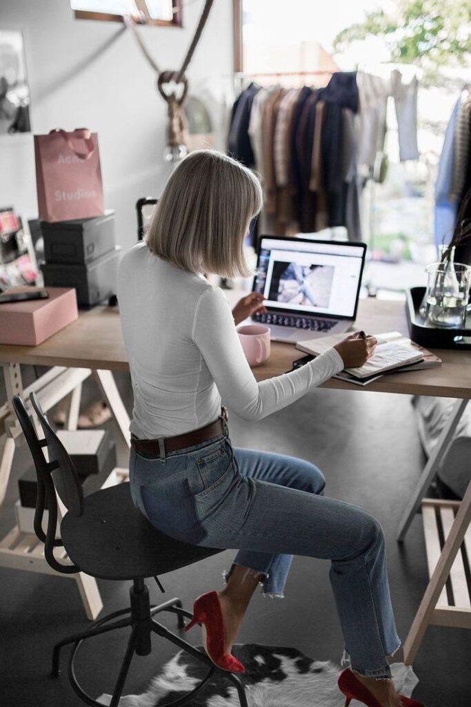 _ 2 Woman working on laptop in stylish home office with clothing rack, seated at desk, wearing jeans and red heels, natural light streaming in. | Sky Rye Design Woman working on laptop in stylish home office with clothing rack, seated at desk, wearing jeans and red heels, natural light streaming in.