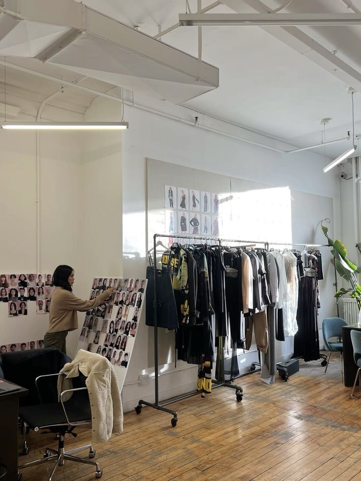 Fashion studio scene with a woman organizing model photos on a board, racks of clothing, and bright natural light filling the room.