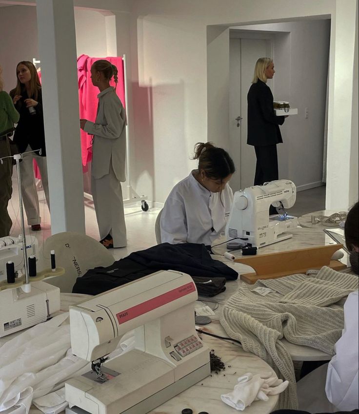 People working on sewing machines in a fashion studio, with fabric and garments around, while others discuss in the background.