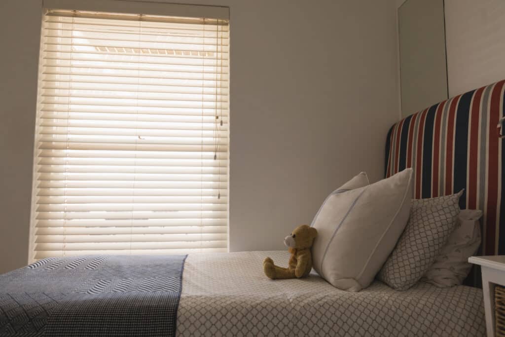Side view of teddy bear on a bed in bedroom at home Cozy bedroom with soft pillows and a teddy bear on a neatly made bed, illuminated by natural light through Venetian blinds. | Sky Rye Design Cozy bedroom with soft pillows and a teddy bear on a neatly made bed, illuminated by natural light through Venetian blinds.