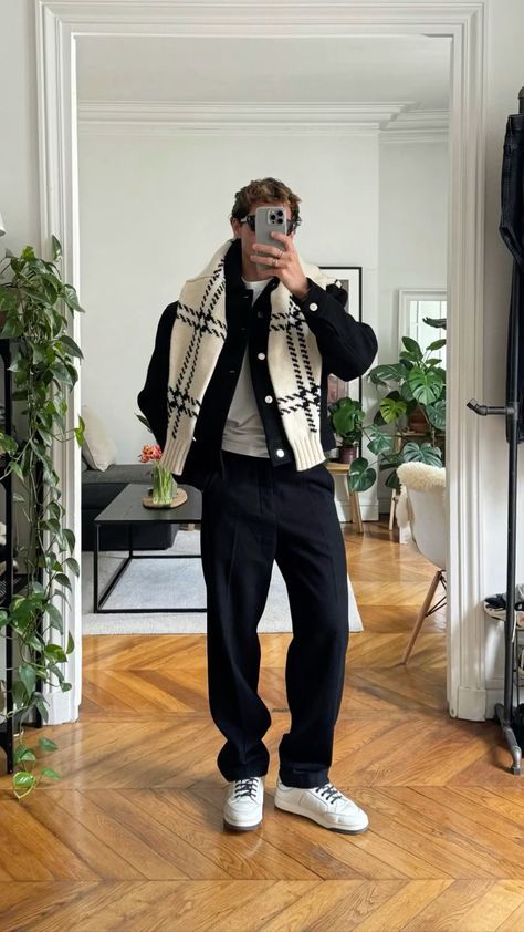 Man in a stylish outfit takes a mirror selfie in a modern living room, wearing a black and white jacket with plants and wood flooring around.