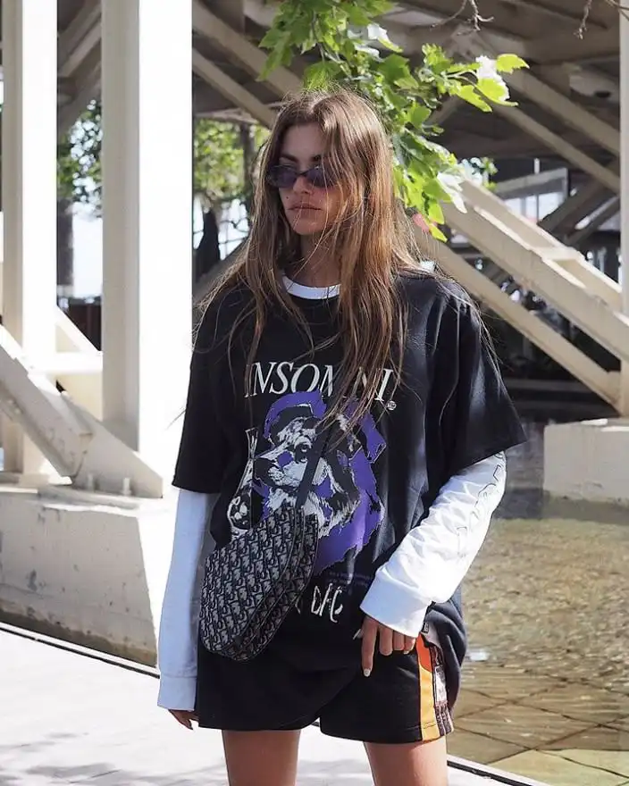 Fashionable woman in layered streetwear outfit with graphic tee, shorts, and sunglasses, standing outdoors by modern architecture.