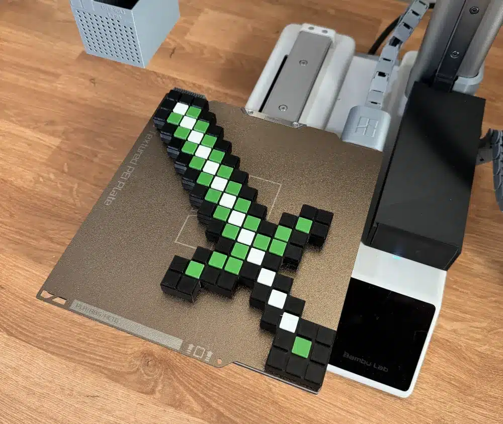 2024-10-26_d559e0eaff9a3 Pixelated sword design with green and black blocks on a 3D printer bed, showcasing a creative 3D printing project on a wood surface. | Sky Rye Design Pixelated sword design with green and black blocks on a 3D printer bed, showcasing a creative 3D printing project on a wood surface.