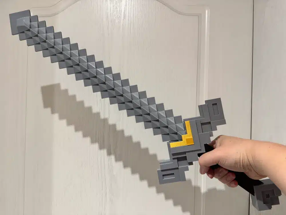 2025-04-05_7cf236b9c14f5 Pixelated toy sword held by hand, casting a shadow on a light wooden door. Retro gaming design with a gray and yellow color scheme. | Sky Rye Design Pixelated toy sword held by hand, casting a shadow on a light wooden door. Retro gaming design with a gray and yellow color scheme.
