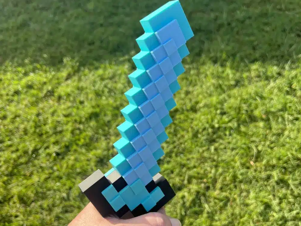 2025-05-12_68300df0f8551 Pixelated toy sword resembling a Minecraft weapon held over grass, showcasing its iconic blue and gray blocky design. | Sky Rye Design minecraft sword 3d print