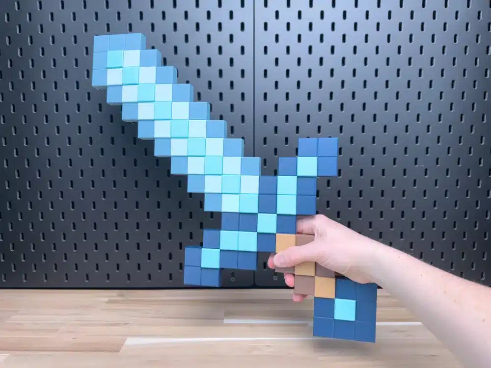 2025-07-13_7d1535170c9368 Hand holding a pixelated blue sword against a perforated black background on a wooden table, resembling a Minecraft design. | Sky Rye Design Hand holding a pixelated blue sword against a perforated black background on a wooden table, resembling a Minecraft design.