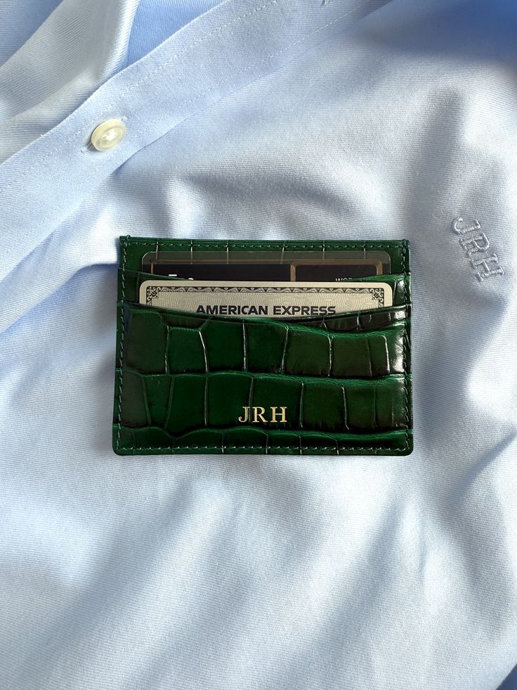 Green leather cardholder with initials JRH holds an American Express card on a light blue shirt background.