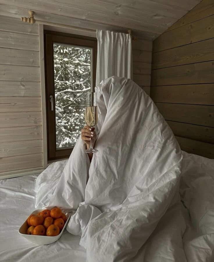 _ - 2025-12-04T130524449 Cozy person under white blanket with champagne, tangerines on bed, snowy window view in rustic cabin setting. | Sky Rye Design Cozy person under white blanket with champagne, tangerines on bed, snowy window view in rustic cabin setting.