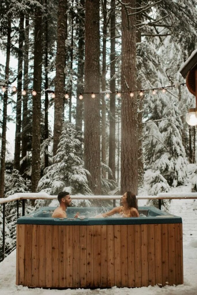 Two people relaxing in an outdoor hot tub surrounded by snow-covered trees and string lights, creating a cozy winter getaway ambiance.