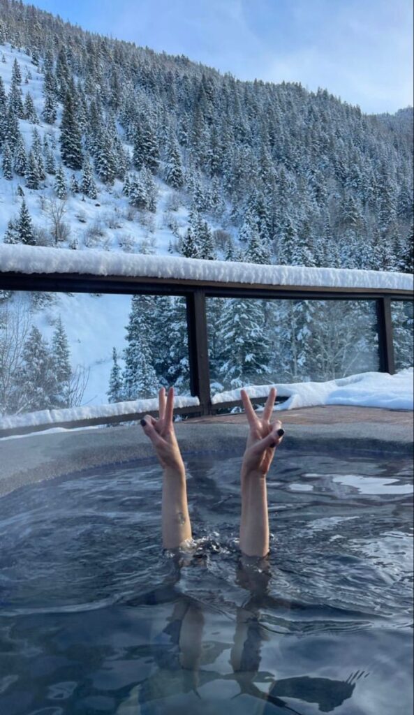 Person enjoying a hot tub in snowy mountains, with hands making peace signs against a winter landscape. Cozy winter relaxation.