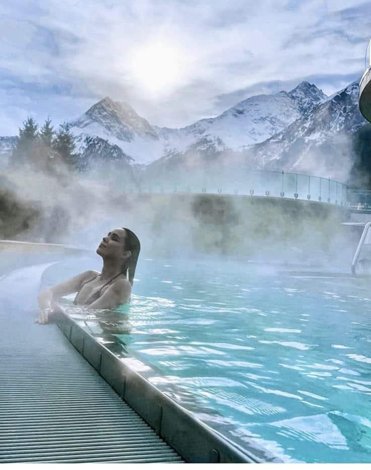 Person relaxing in a steamy outdoor pool with scenic snowy mountains and a bright sun in the background.