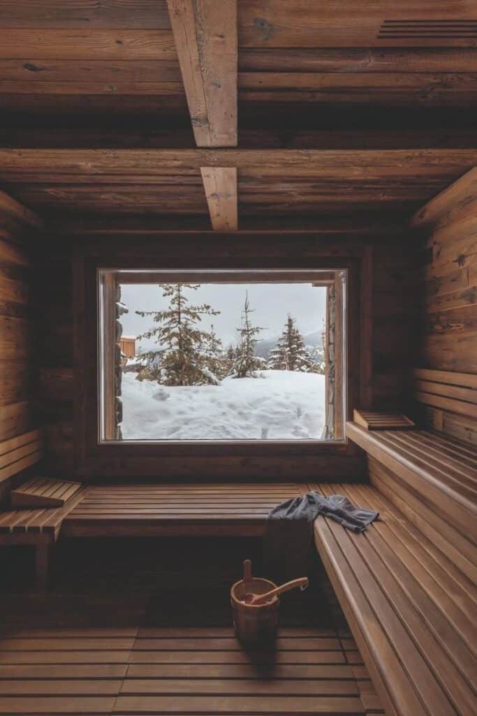 Cozy wooden sauna interior with snowy mountain view through large window, featuring bench seating and a sauna bucket on the floor.