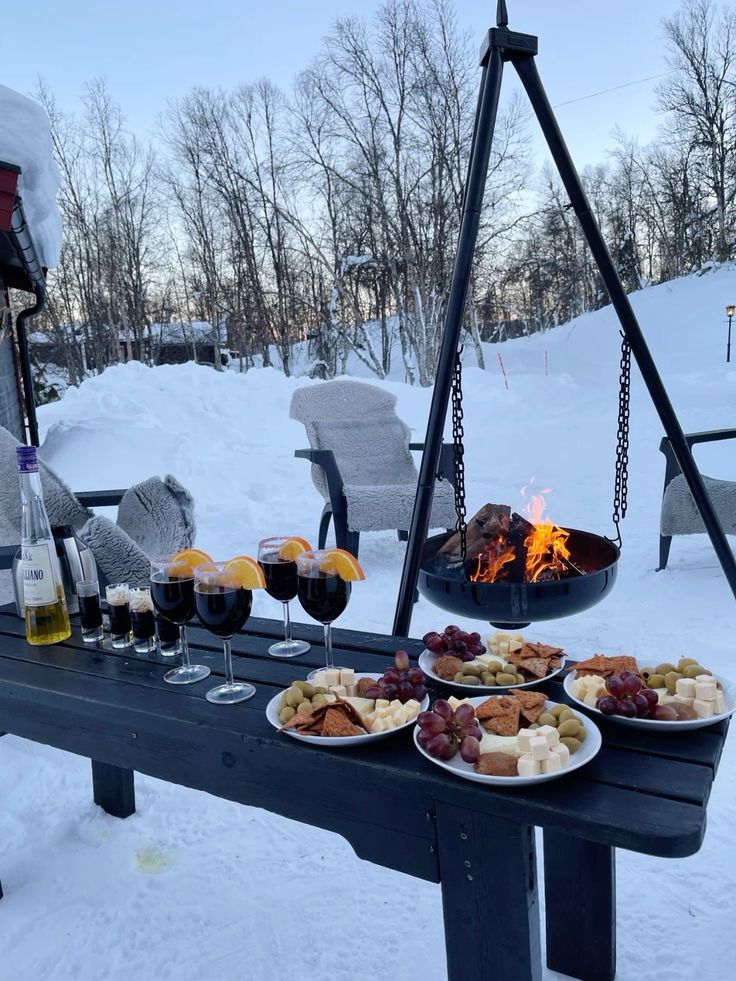 Outdoor winter picnic with snacks, red wine, and a cozy fire in the snow, surrounded by snowy trees and chairs with blankets.