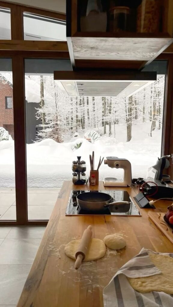 Modern kitchen with dough rolling on a wooden countertop, snowy winter view through large windows in the background.