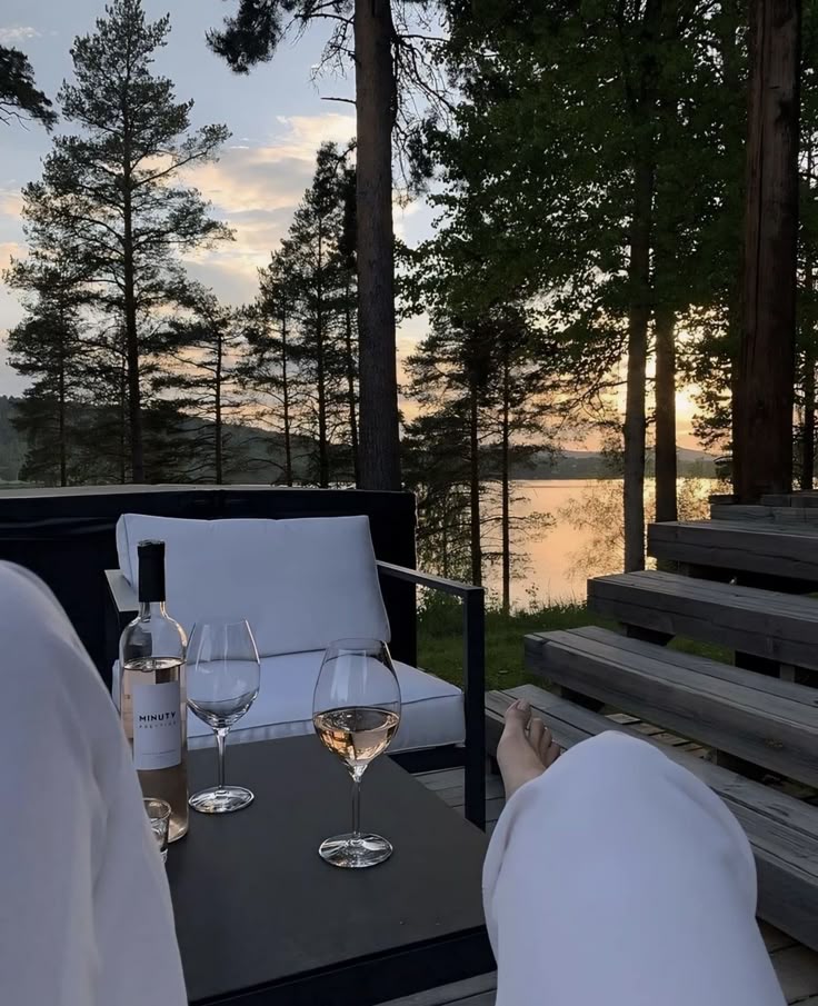 Screenshot Relaxing lakeside evening, two wine glasses and white furniture on a deck surrounded by trees at sunset. | Sky Rye Design Relaxing lakeside evening, two wine glasses and white furniture on a deck surrounded by trees at sunset.