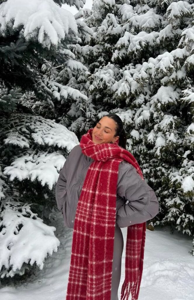 Person smiling in snowy forest, wearing a red plaid scarf and gray jacket, enjoying winter wonderland. Cozy and festive winter scene.