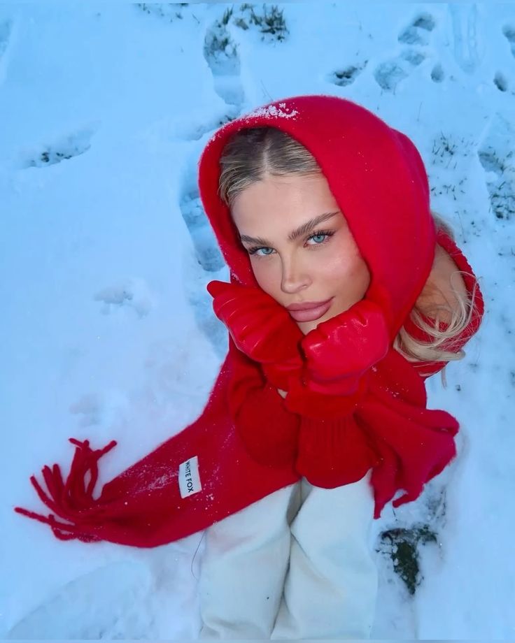 Woman in red scarf and gloves sitting in snow, looking up. Cozy winter attire, vibrant red contrast with white background.