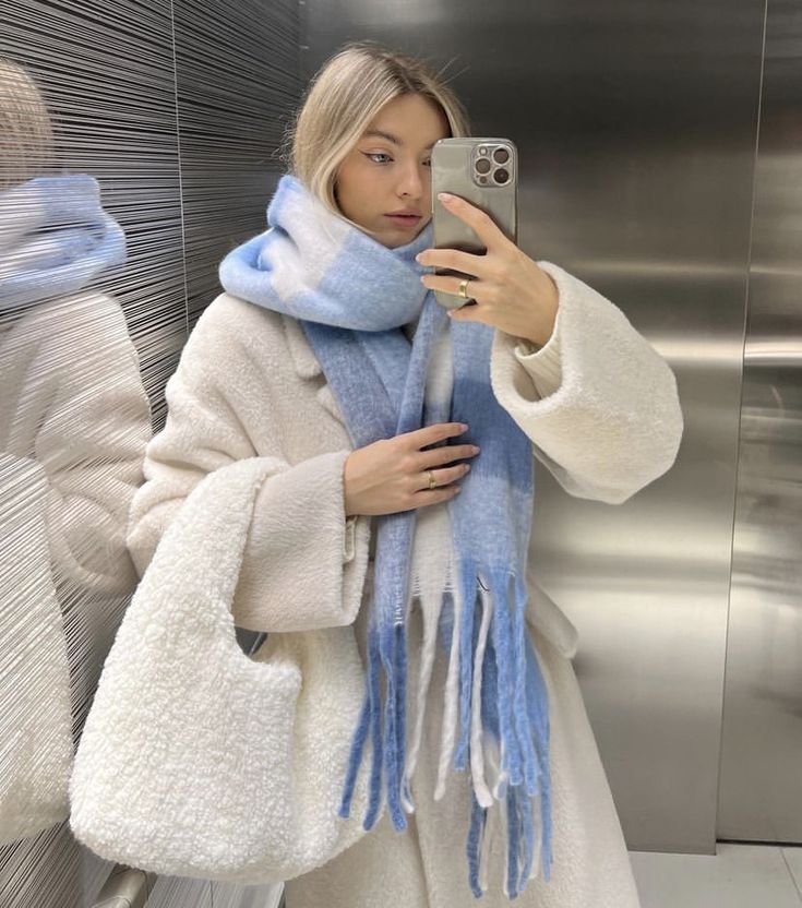 Woman in a mirror selfie, wearing a white coat and blue scarf, holding a matching white bag, captures a chic winter fashion look.