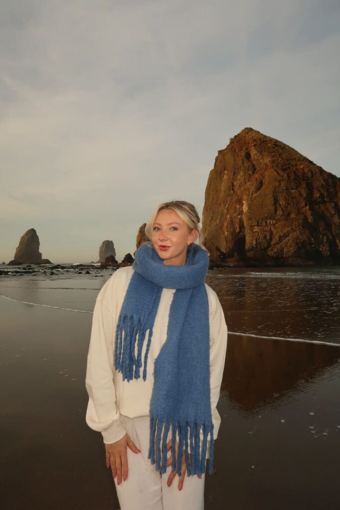 Person wearing a blue scarf on a beach by large rock formations at sunset, creating a serene and picturesque coastal scene.
