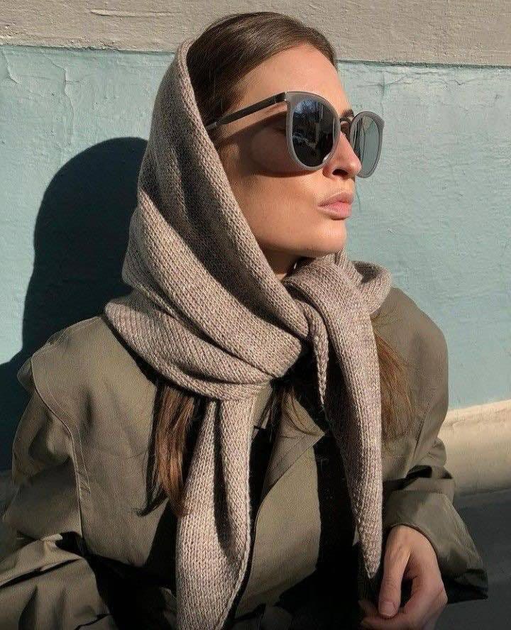 Woman in sunglasses and coat with a knitted scarf, standing against a sunlit wall.