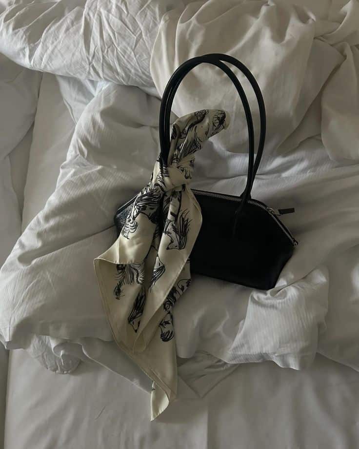 Black handbag with a patterned scarf placed on white rumpled bed sheets, creating a chic and cozy aesthetic.