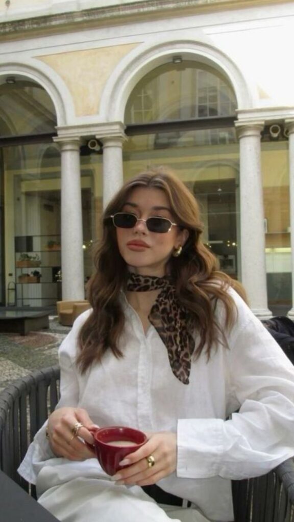 Woman in white shirt and sunglasses sipping coffee at an outdoor café, wearing a leopard-print scarf and gold earrings.