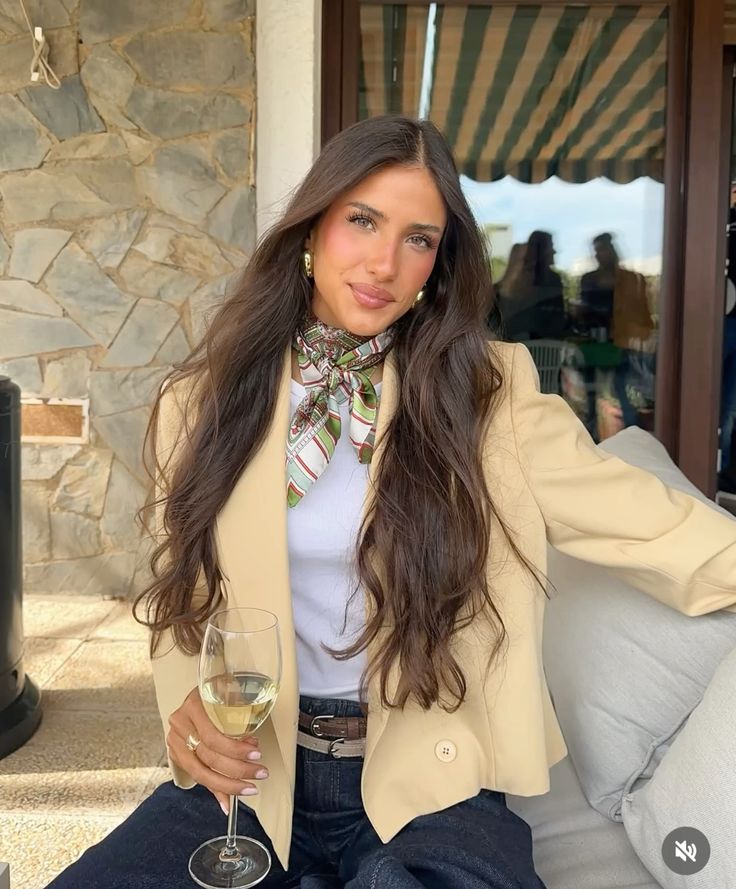 Stylish woman in beige blazer and scarf enjoying a drink outdoors, seated on a patio with a stone wall background.