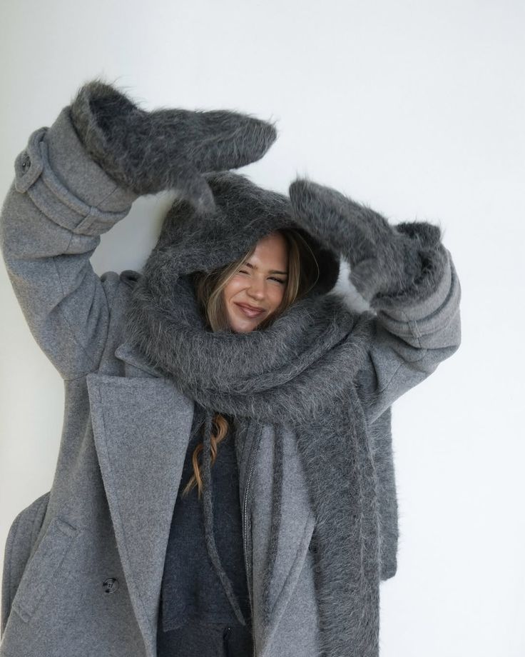 Smiling woman in oversized gray coat and fluffy scarf, playfully posing with raised arms, creating a cozy winter fashion vibe.
