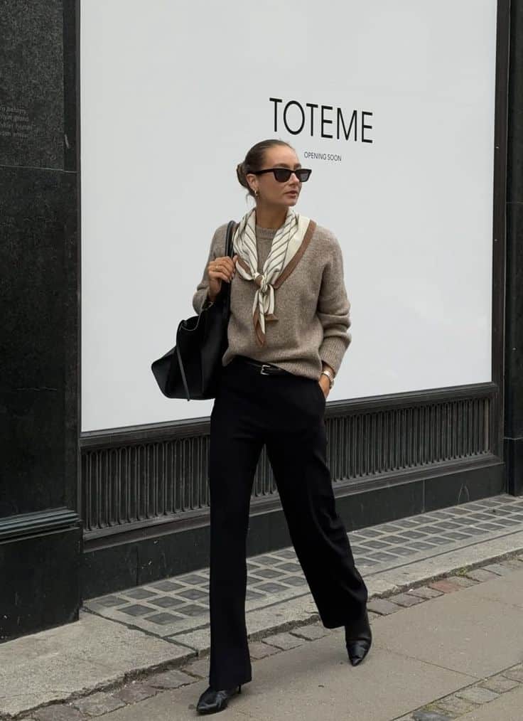 Fashionable woman in chic outfit with scarf and sunglasses walks by Toteme store opening soon. Elegant street style fashion.