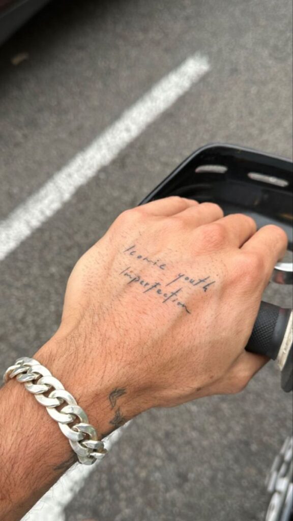 Close-up of a hand on a bicycle handlebar with a tattoo reading Iconic youth imperfection and a silver chain bracelet.