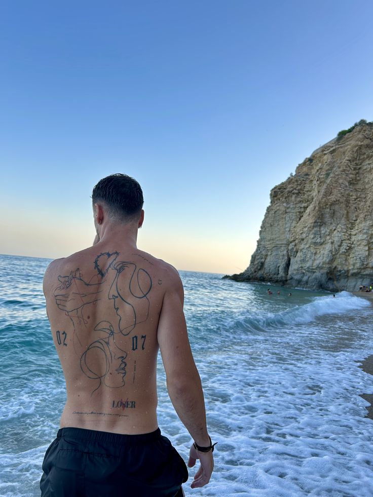 Man with back tattoos walking along a scenic beach with waves and towering cliffs at sunset, capturing a serene summer moment.
