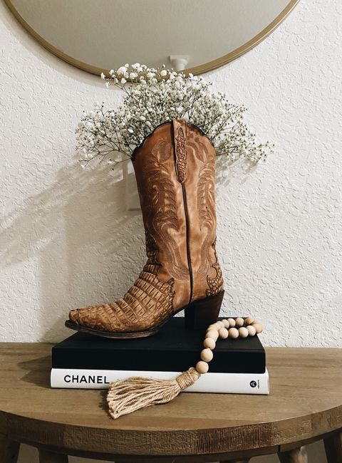 _ - 2025-12-10T134045479 Cowboy boot vase with flowers atop Chanel books on a wooden table, accented with a beaded tassel, in front of a circular mirror. | Sky Rye Design Cowboy boot vase with flowers atop Chanel books on a wooden table, accented with a beaded tassel, in front of a circular mirror.
