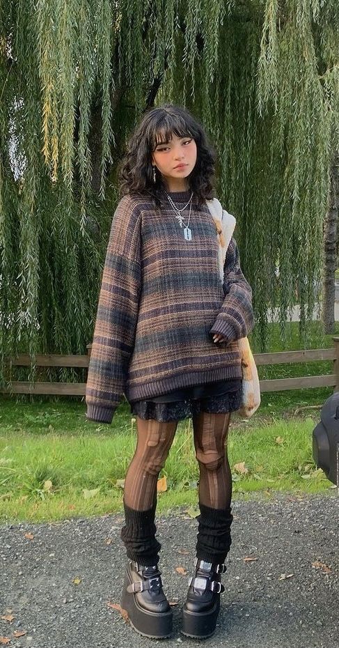 Person in a plaid sweater with layered accessories, standing on a path in front of trees, showcasing a unique, eclectic fashion style.