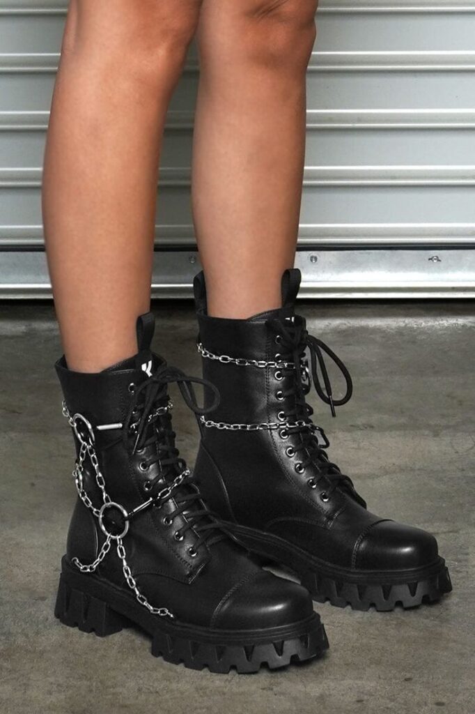 Black leather combat boots with chain details, worn indoors on a concrete floor, offering a bold statement look.