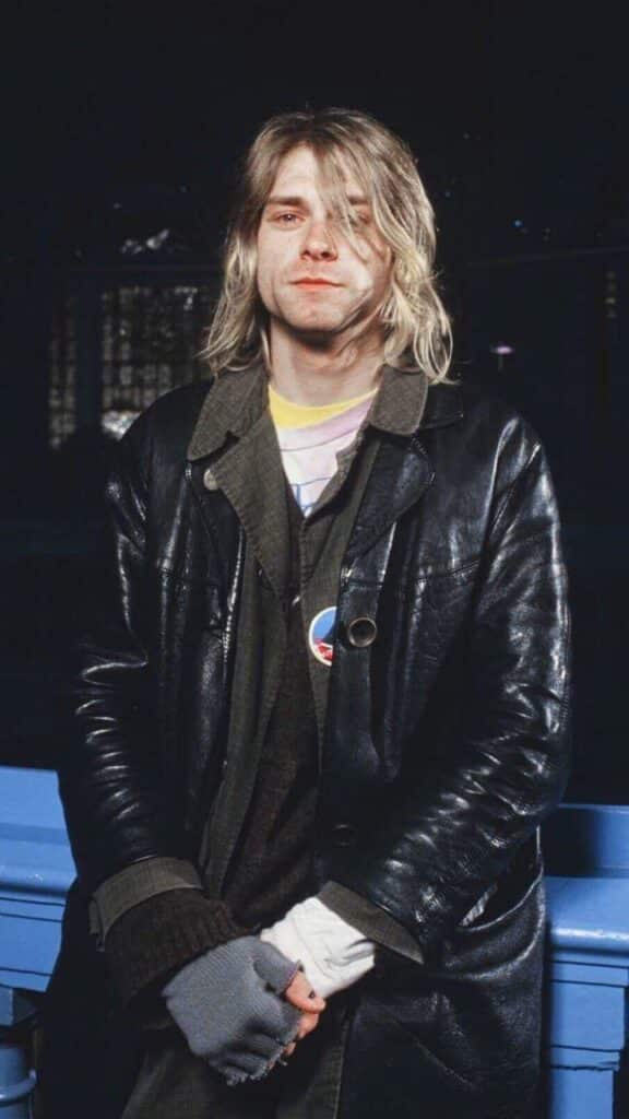 Young man in a leather jacket and gloves, standing indoors with long blonde hair, looking at the camera with a neutral expression.