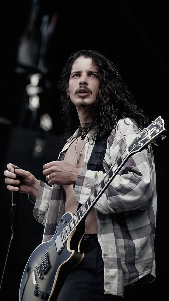 Long-haired musician with a guitar performs on stage, wearing an open plaid shirt, capturing an intense moment of live music energy.