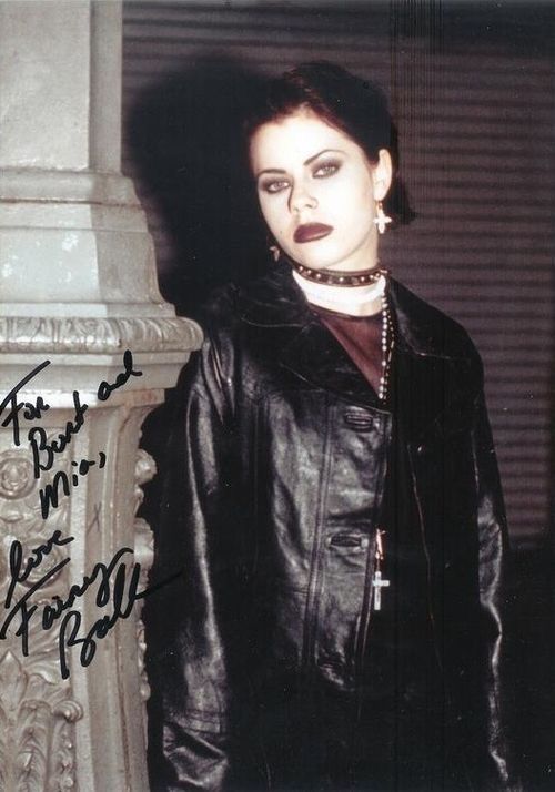 A person in goth attire with dark makeup and a choker stands near a pillar, wearing a leather jacket and cross earrings, gazing at the camera.